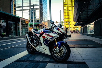 A motorcycle with a sporty design parked on a modern city street, surrounded by tall buildings with reflective glass surfaces. The environment shows a blend of urban architecture and outdoor elements, with a person in the background near a storefront.