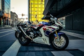 A sport motorcycle with a sleek design is parked on a modern city street. The bike features a striking color scheme of white, red, and blue, and the 'Honda' logo is visible on its side. In the background, contemporary buildings with reflective glass panes and a bright yellow structure add a dynamic urban feel.