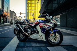 A sport motorcycle with a sleek design is parked on a modern city street. The bike features a striking color scheme of white, red, and blue, and the 'Honda' logo is visible on its side. In the background, contemporary buildings with reflective glass panes and a bright yellow structure add a dynamic urban feel.