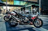 Three sport motorcycles are parked on an urban street. The motorcycles feature sleek designs with dominant colors of black, red, white, and blue. They are positioned side by side on a neatly paved area, with city buildings and a glass walkway visible in the background.