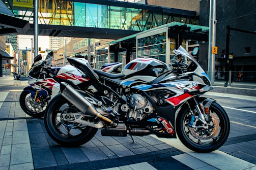 Three sport motorcycles are parked on an urban street. The motorcycles feature sleek designs with dominant colors of black, red, white, and blue. They are positioned side by side on a neatly paved area, with city buildings and a glass walkway visible in the background.