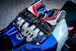 A pair of multi-colored, armored motorcycle gloves rest on a sleek, blue and white sport bike seat. The gloves feature intricate designs with dominant colors of blue, red, white, and black. Surrounding the gloves is textured material, and part of the bike's branding and design is visible.