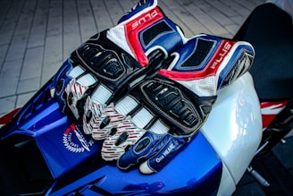 A pair of multi-colored, armored motorcycle gloves rest on a sleek, blue and white sport bike seat. The gloves feature intricate designs with dominant colors of blue, red, white, and black. Surrounding the gloves is textured material, and part of the bike's branding and design is visible.