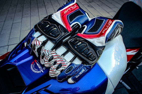 A pair of multi-colored, armored motorcycle gloves rest on a sleek, blue and white sport bike seat. The gloves feature intricate designs with dominant colors of blue, red, white, and black. Surrounding the gloves is textured material, and part of the bike's branding and design is visible.