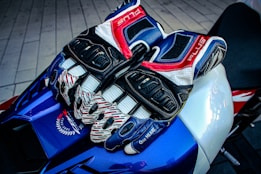 A pair of multi-colored, armored motorcycle gloves rest on a sleek, blue and white sport bike seat. The gloves feature intricate designs with dominant colors of blue, red, white, and black. Surrounding the gloves is textured material, and part of the bike's branding and design is visible.