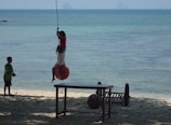 Children are playing on a beach by the ocean, with one child swinging on a rope attached to a large red buoy. Another child watches from the sand nearby. A table and some items are on the sand.
