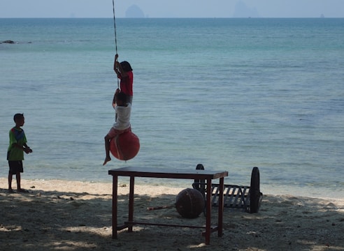 Children are playing on a beach by the ocean, with one child swinging on a rope attached to a large red buoy. Another child watches from the sand nearby. A table and some items are on the sand.