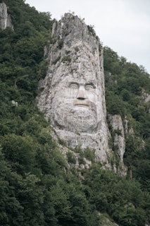 A large rock carving featuring the face of a man with a determined expression is carved into a rugged cliff. The surrounding landscape is dense with green foliage, creating a natural frame for the imposing sculpture. Inscribed below the face are the words 'Decebalus Rex Dragan Fecit'. At the top of the rocky formation is a small cross.