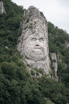 A large rock carving featuring the face of a man with a determined expression is carved into a rugged cliff. The surrounding landscape is dense with green foliage, creating a natural frame for the imposing sculpture. Inscribed below the face are the words 'Decebalus Rex Dragan Fecit'. At the top of the rocky formation is a small cross.