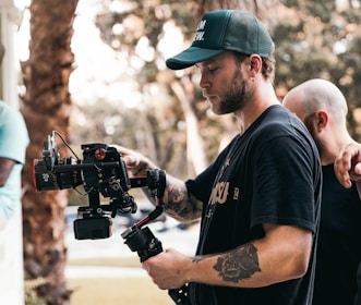 A man with tattoos is operating a professional video camera on a stabilizer. He is wearing a green cap and a black t-shirt. Another person with a shaved head is partially visible in the background, placing a hand on his shoulder. The setting appears to be outdoors, with blurred trees and sunlight creating a soft bokeh effect in the background.