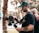 A man with tattoos is operating a professional video camera on a stabilizer. He is wearing a green cap and a black t-shirt. Another person with a shaved head is partially visible in the background, placing a hand on his shoulder. The setting appears to be outdoors, with blurred trees and sunlight creating a soft bokeh effect in the background.