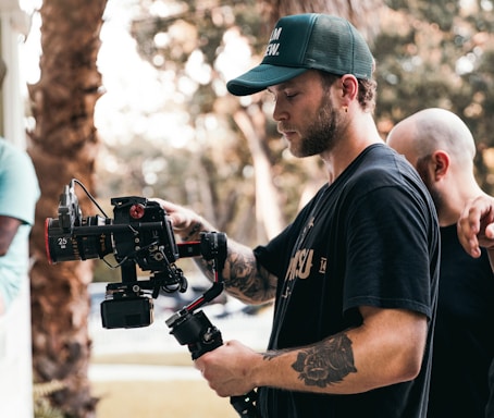 A man with tattoos is operating a professional video camera on a stabilizer. He is wearing a green cap and a black t-shirt. Another person with a shaved head is partially visible in the background, placing a hand on his shoulder. The setting appears to be outdoors, with blurred trees and sunlight creating a soft bokeh effect in the background.