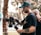 A man with tattoos is operating a professional video camera on a stabilizer. He is wearing a green cap and a black t-shirt. Another person with a shaved head is partially visible in the background, placing a hand on his shoulder. The setting appears to be outdoors, with blurred trees and sunlight creating a soft bokeh effect in the background.