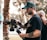A man with tattoos is operating a professional video camera on a stabilizer. He is wearing a green cap and a black t-shirt. Another person with a shaved head is partially visible in the background, placing a hand on his shoulder. The setting appears to be outdoors, with blurred trees and sunlight creating a soft bokeh effect in the background.