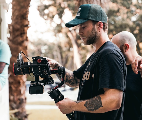 A man with tattoos is operating a professional video camera on a stabilizer. He is wearing a green cap and a black t-shirt. Another person with a shaved head is partially visible in the background, placing a hand on his shoulder. The setting appears to be outdoors, with blurred trees and sunlight creating a soft bokeh effect in the background.