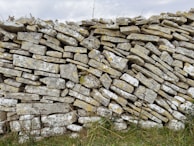 A dry stone wall constructed from irregularly shaped stones stacked on top of each other. The stones have a rough, weathered texture and display various shades of gray and beige, with patches of moss and lichen adding a hint of yellow. Some grasses and small plants grow at the base of the wall, suggesting it is situated in a natural or rural setting.