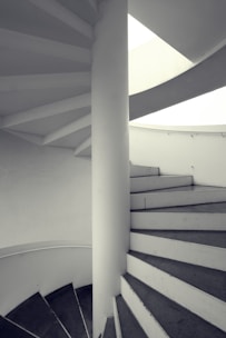 A spiral staircase with a central supporting pillar, featuring clean, white steps and a minimalist design. The stairs curve upwards and are bathed in natural light from an opening above.