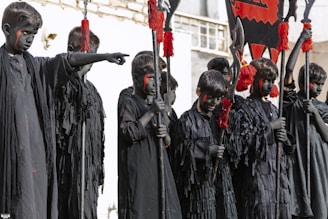A group of children dressed in black robes with black face paint stand together, each holding a pole adorned with red tassels and flags. The children exhibit a serious demeanor, and their faces are painted with red markings.