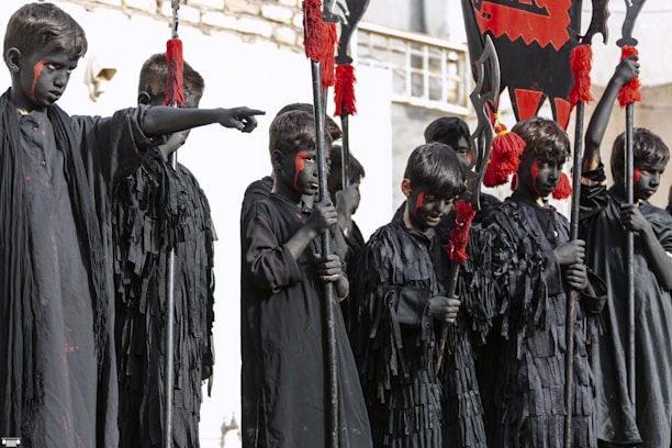 A group of children dressed in black robes with black face paint stand together, each holding a pole adorned with red tassels and flags. The children exhibit a serious demeanor, and their faces are painted with red markings.