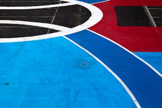 A geometric and abstract design featuring large, intersecting shapes in vibrant colors. Thick white lines create curved patterns over expansive areas of bright blue, red, and black. The composition is symmetrical and bold, with a sense of dynamic movement.
