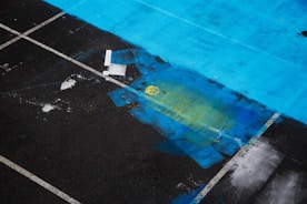 Different sections of a parking lot with blue, yellow, and black areas can be seen, along with white parking lines. There is some abstract paint on the ground, including rectangular shapes and patches of color.