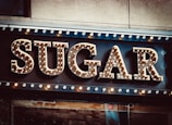Large marquee-style letters spelling out the word 'SUGAR' with light bulbs decorating each letter, creating a vintage or classic sign appearance. The background is a dark surface with additional lights bordering the top.