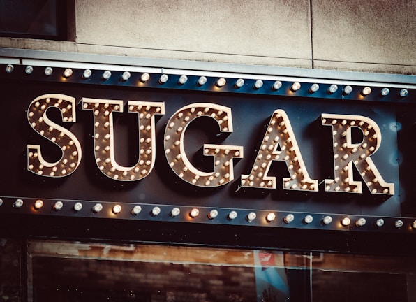 Large marquee-style letters spelling out the word 'SUGAR' with light bulbs decorating each letter, creating a vintage or classic sign appearance. The background is a dark surface with additional lights bordering the top.