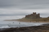 A large medieval castle sits atop a hill overlooking a sandy beach, with waves gently crashing onto the shore. Several people in wetsuits are visible on surfboards, while others walk along the beach, bundled up against the overcast weather. The sky is filled with gray clouds, adding a moody atmosphere to the scene.