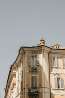 A classic European-style building with ornate architectural details, including decorative elements around windows and wrought iron balconies. The façade of the building is painted in light earthy tones, and some windows have shutters. The sky above is clear and blue, adding to the serene atmosphere of the scene.