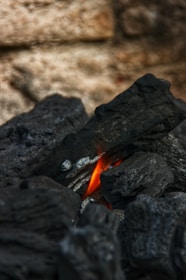 Dark charcoal pieces are stacked on top of each other, with an orange and red flame flickering between them. The background is blurred, giving focus to the charcoal and fire.