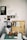 An art workspace featuring a wooden box open with various art supplies inside, including paintbrushes and printmaking tools. Two prints of leaves are clipped on the lid of the box. A green cutting mat is on the table, with a glass jar and paper towels. To the side, a lamp stands with a pair of black headphones hanging on it. The wall is adorned with sketches and photographs.