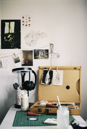An art workspace featuring a wooden box open with various art supplies inside, including paintbrushes and printmaking tools. Two prints of leaves are clipped on the lid of the box. A green cutting mat is on the table, with a glass jar and paper towels. To the side, a lamp stands with a pair of black headphones hanging on it. The wall is adorned with sketches and photographs.