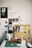 An art workspace featuring a wooden box open with various art supplies inside, including paintbrushes and printmaking tools. Two prints of leaves are clipped on the lid of the box. A green cutting mat is on the table, with a glass jar and paper towels. To the side, a lamp stands with a pair of black headphones hanging on it. The wall is adorned with sketches and photographs.