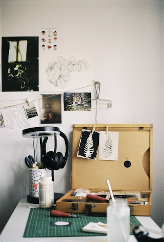 An art workspace featuring a wooden box open with various art supplies inside, including paintbrushes and printmaking tools. Two prints of leaves are clipped on the lid of the box. A green cutting mat is on the table, with a glass jar and paper towels. To the side, a lamp stands with a pair of black headphones hanging on it. The wall is adorned with sketches and photographs.