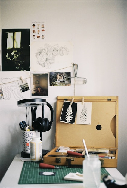 An art workspace featuring a wooden box open with various art supplies inside, including paintbrushes and printmaking tools. Two prints of leaves are clipped on the lid of the box. A green cutting mat is on the table, with a glass jar and paper towels. To the side, a lamp stands with a pair of black headphones hanging on it. The wall is adorned with sketches and photographs.