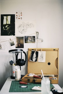 An art workspace featuring a wooden box open with various art supplies inside, including paintbrushes and printmaking tools. Two prints of leaves are clipped on the lid of the box. A green cutting mat is on the table, with a glass jar and paper towels. To the side, a lamp stands with a pair of black headphones hanging on it. The wall is adorned with sketches and photographs.