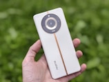A hand is holding a sleek white smartphone with a circular camera module. The phone has a light orange vertical strip running through the center. The brand name 'realme' is visible on its back. The background is blurred, consisting of lush green foliage.