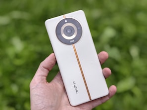 A hand is holding a sleek white smartphone with a circular camera module. The phone has a light orange vertical strip running through the center. The brand name 'realme' is visible on its back. The background is blurred, consisting of lush green foliage.