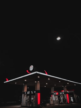 A gas station is illuminated at night under a dark sky. The station's canopy and fuel pumps are lit, casting a soft glow in the surrounding darkness. The canopy features distinct red and white branding, and a single bright streetlamp provides additional lighting from above.