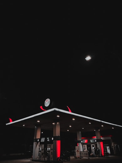 A gas station is illuminated at night under a dark sky. The station's canopy and fuel pumps are lit, casting a soft glow in the surrounding darkness. The canopy features distinct red and white branding, and a single bright streetlamp provides additional lighting from above.