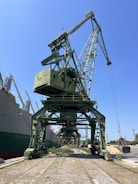 A large green industrial crane is positioned at a dockyard, towering over the surrounding area. It is situated next to a series of large ships, with a clear blue sky overhead. The area around the crane shows vegetation growing between the cobblestone path, indicating minimal activity or maintenance.