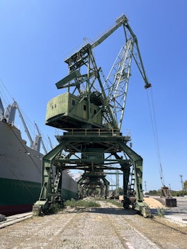 A large green industrial crane is positioned at a dockyard, towering over the surrounding area. It is situated next to a series of large ships, with a clear blue sky overhead. The area around the crane shows vegetation growing between the cobblestone path, indicating minimal activity or maintenance.
