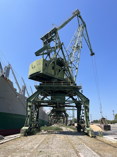 A large green industrial crane is positioned at a dockyard, towering over the surrounding area. It is situated next to a series of large ships, with a clear blue sky overhead. The area around the crane shows vegetation growing between the cobblestone path, indicating minimal activity or maintenance.