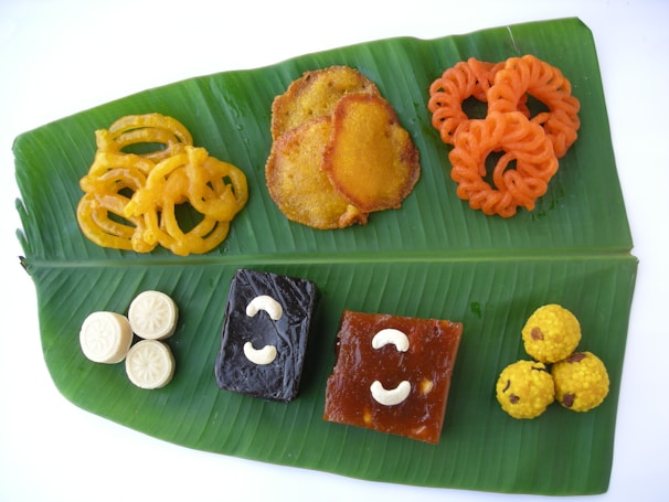 A variety of colorful Indian sweets are arranged neatly on a large green banana leaf. The sweets include swirling yellow and orange jalebis, round yellow laddoos, dark square pieces with cashew nuts, and white round shapes resembling peda or sandesh. The arrangement on the banana leaf highlights the traditional presentation style.