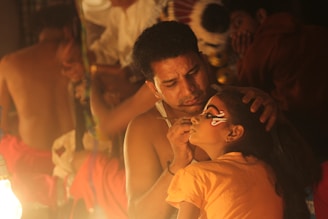 A man is carefully applying intricate face paint to a young girl in a dimly lit setting. They appear to be preparing for a cultural or theatrical performance. The focus is on the artistry and concentration required for the task.