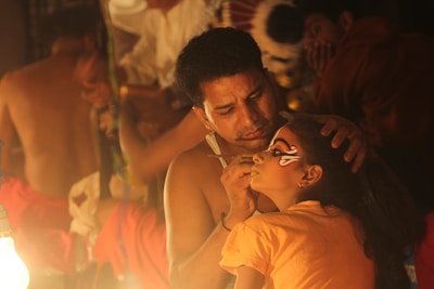 A man is carefully applying intricate face paint to a young girl in a dimly lit setting. They appear to be preparing for a cultural or theatrical performance. The focus is on the artistry and concentration required for the task.