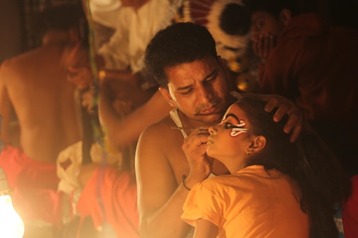 A man is carefully applying intricate face paint to a young girl in a dimly lit setting. They appear to be preparing for a cultural or theatrical performance. The focus is on the artistry and concentration required for the task.