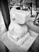 A stone sculpture with a geometric, abstract form sits on a wooden stand. Surrounding the main sculpture are several other sculptures at different stages of completion, placed on wooden trestles in a workshop setting. The environment appears orderly, with tools and materials organized around.
