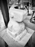 A stone sculpture with a geometric, abstract form sits on a wooden stand. Surrounding the main sculpture are several other sculptures at different stages of completion, placed on wooden trestles in a workshop setting. The environment appears orderly, with tools and materials organized around.
