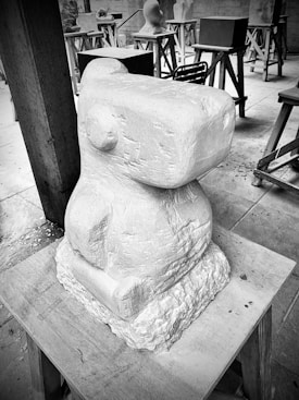 A stone sculpture with a geometric, abstract form sits on a wooden stand. Surrounding the main sculpture are several other sculptures at different stages of completion, placed on wooden trestles in a workshop setting. The environment appears orderly, with tools and materials organized around.